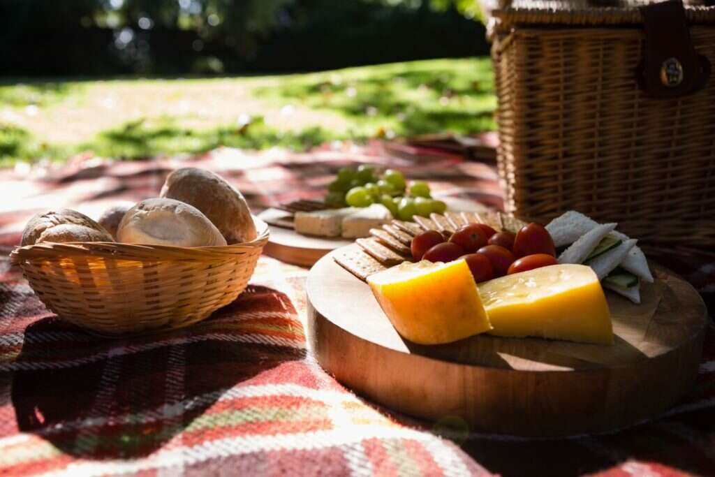 Slika hrane na pikniku. Na slici se nalazi sir, paradajz, korpa za piknik.